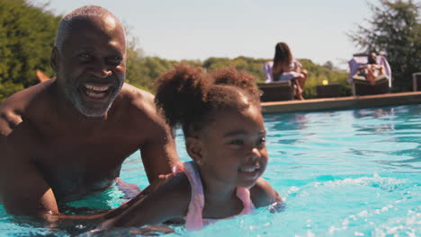 Grandfather-Teaching-Granddaughter-To-Swim-In-Outdoor-Pool-On-Holiday