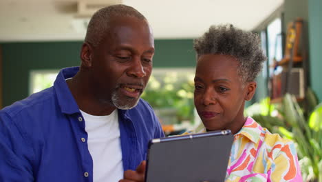 Lächelndes-älteres-Paar,-Das-Zu-Hause-Sitzt-Und-Einen-Urlaub-Bucht-Oder-Online-Auf-Einem-Digitalen-Tablet-Einkauft