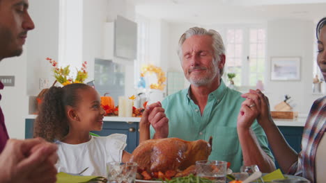 Grandfather-Talking-To-Granddaughter-As-Multi-Generation-Family-Pray-Before-Thanksgiving-Meal