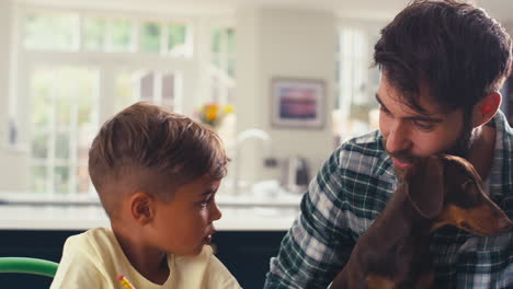Father-Holding-Pet-Dachshund-At-Home-In-Kitchen-Helping-Son-With-Homework