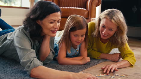 Same-Sex-Family-With-Two-Mature-Mums-And-Daughter-Lying-On-Floor-Doing-Jigsaw-Puzzle-At-Home