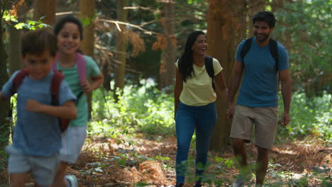 Family-With-Backpacks-Hiking-Or-Walking-In-Summer-Woodland-Countryside-With-Children-Running-Ahead