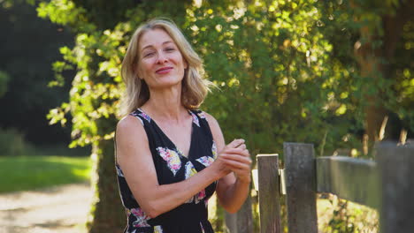 Portrait-Of-Casually-Dressed-Mature-Woman-Leaning-On-Fence-On-Walk-In-Countryside