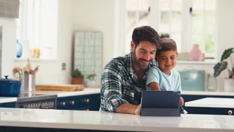Vater-Zu-Hause-In-Der-Küche-Mit-Sohn,-Der-Auf-Einem-Digitalen-Tablet-Streamt-Oder-Ein-Spiel-Spielt