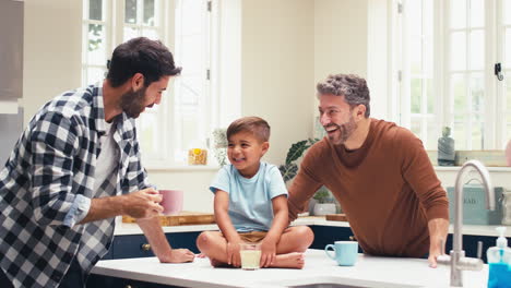 Same-Sex-Family-With-Two-Dads-In-Kitchen-With-Son-Sitting-On-Counter