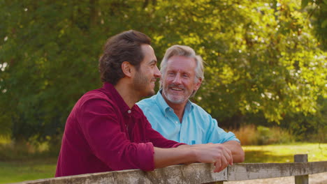 Mature-Father-With-Mixed-Race-Adult-Son-Leaning-On-Fence-Walking-In-Countryside
