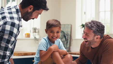 Same-Sex-Family-With-Two-Dads-In-Kitchen-Tickling-Son-Sitting-On-Counter
