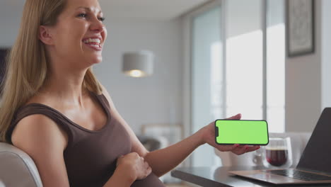 Mujer-Embarazada-Sonriente-En-Casa-Trabajando-En-Una-Computadora-Portátil-Sosteniendo-Un-Teléfono-Móvil-De-Pantalla-Verde