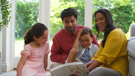 Lächelnde-Familie-Zu-Hause-Sitzt-Auf-Dem-Boden-Im-Wohnzimmer-Und-Liest-Gemeinsam-Ein-Buch