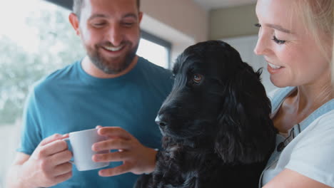 Una-Pareja-Bebiendo-Café-En-La-Cocina-De-Casa-Jugando-Con-Un-Perro-Spaniel-Como-Mascota