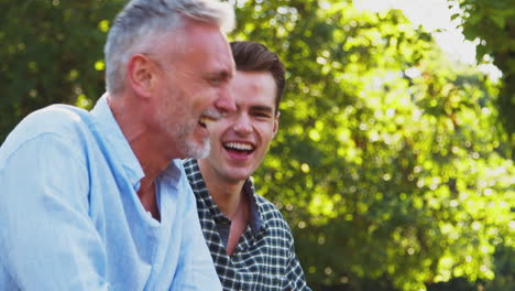 Mature-Father-With-Adult-Son-Leaning-On-Fence-And-Laughing-Walking-In-Countryside