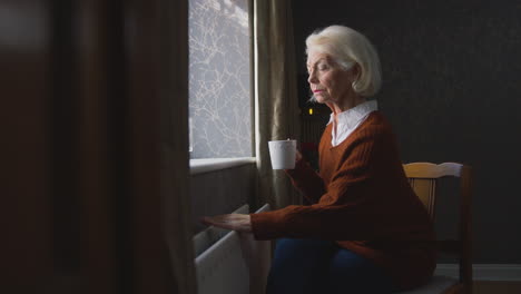 Senior-Woman-With-Hot-Drink-Trying-To-Keep-Warm-By-Radiator-At-Home-In-Cost-Of-Living-Energy-Crisis