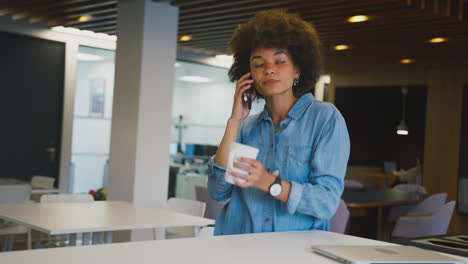 Businesswoman-Standing-In-Modern-Open-Plan-Office-Talking-On-Mobile-Phone