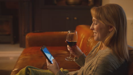 Frau-Sitzt-Zu-Hause-Auf-Dem-Sofa-Mit-Feuer-Und-Hat-Ein-Handy-Mit-Blauem-Bildschirm,-Das-Ein-Glas-Wein-Hält