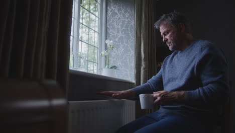 Unhappy-Mature-Man-Trying-To-Keep-Warm-By-Radiator-At-Home-During-Cost-Of-Living-Energy-Crisis