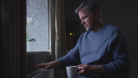 Unhappy-Mature-Man-Trying-To-Keep-Warm-By-Radiator-At-Home-During-Cost-Of-Living-Energy-Crisis