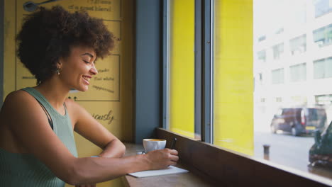Female-Customer-In-Coffee-Shop-Window-Working-Writing-In-Notebook