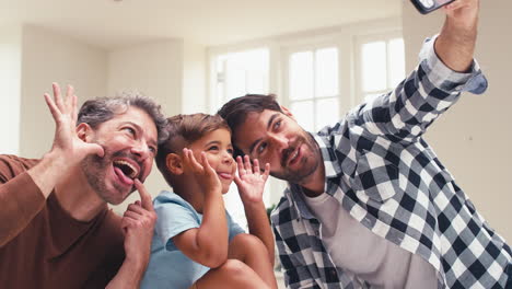 Gleichgeschlechtliche-Familie-Mit-Zwei-Vätern,-Die-Grimassen-Für-Ein-Selfie-In-Der-Küche-Ziehen,-Während-Ihr-Sohn-Auf-Der-Theke-Sitzt