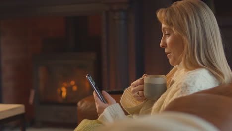 Woman-At-Home-In-Lounge-With-Cosy-Fire-And-Hot-Drink-Using-Mobile-Phone