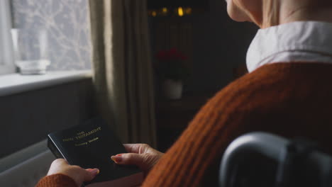 Senior-Woman-In-Wheelchair-With-Bible-Keeping-Warm-By-Radiator-At-Home-In-Cost-Of-Living-Crisis