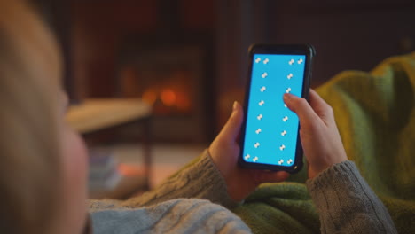 Woman-At-Home-Lying-On-Sofa-In-Lounge-With-Cosy-Fire-Using-Blue-Screen-Mobile-Phone
