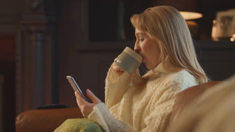 Woman-At-Home-In-Lounge-With-Cosy-Fire-And-Hot-Drink-Using-Mobile-Phone