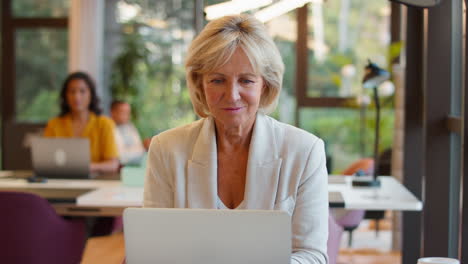 Mature-Businesswoman-Working-On-Laptop-At-Desk-In-Office-Pausing-To-Look-Out-Of-Window