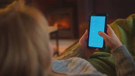 Woman-At-Home-Lying-On-Sofa-In-Lounge-With-Cosy-Fire-Using-Blue-Screen-Mobile-Phone