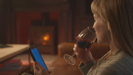Woman-At-Home-Sitting-On-Sofa-With-Fire-With-Blue-Screen-Mobile-Phone-Holding-Glass-Of-Wine