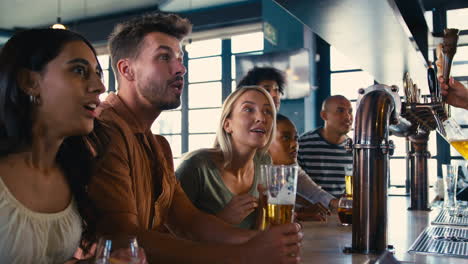 Multi-Cultural-Group-Of-Friends-In-Sports-Bar-Celebrating-As-They-Watch-Game-On-TV