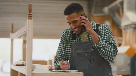 Male-Apprentice-Working-As-Carpenter-In-Furniture-Workshop-Talking-On-Mobile-Phone