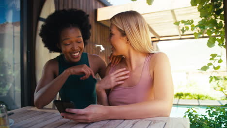 Two-Smiling-Multi-Cultural-Female-Friends-Outdoors-At-Home-Looking-At-Mobile-Phone