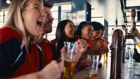 Multi-Cultural-Group-Of-Friends-Wearing-Team-Shirts-In-Sports-Bar-Celebrating-Watching-Game-On-TV