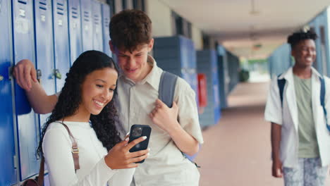 Two-High-School-Or-Secondary-Students-Looking-At-Social-Media-Or-Internet-On-Phone-By-Lockers