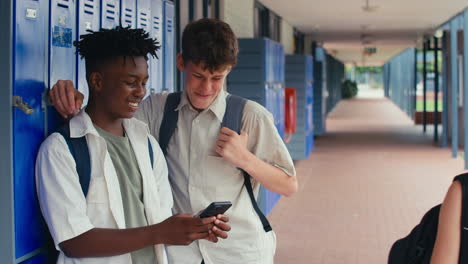 Two-Male-High-School-Or-Secondary-Students-Looking-At-Social-Media-Or-Internet-On-Phone-By-Lockers