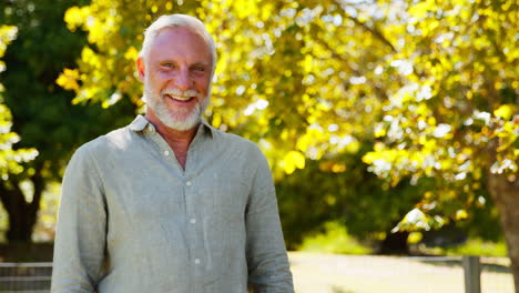 Portrait-Of-Smiling-Senior-Man-Standing-Outdoors-In-Garden-Park-Or-Countryside