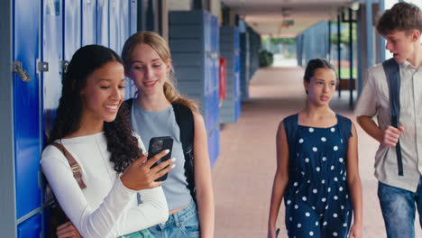 Two-Female-High-School-Or-Secondary-Students-Looking-At-Social-Media-Or-Internet-On-Phone-By-Lockers