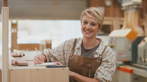 Portrait-Of-Smiling-Female-Apprentice-Carpenter-Working-In-Furniture-Workshop-Making-Notes