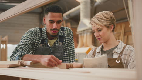 Male-And-Female-Apprentices-Working-As-Carpenters-In-Furniture-Workshop-Measure-Wood-And-Take-Notes