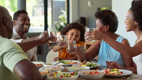 Mehrgenerationenfamilie-Am-Tisch,-Die-Mit-Wasser-Jubelt,-Bevor-Sie-Zu-Hause-Essen-Zum-Essen-Serviert