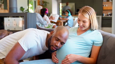 Couple-With-Man-Listening-To-Pregnant-Woman's-Stomach-On-Sofa-At-Home-With-Multi-Generation-Family