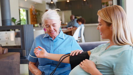 Ältere-Hebamme-Besucht-Schwangere-Frau-Zu-Hause-Und-Misst-Mit-Der-Familie-Im-Hintergrund-Den-Blutdruck