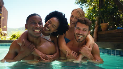 Portrait-Of-Multi-Cultural-Friends-On-Holiday-In-Swimming-Pool-With-Men-Giving-Women-Piggybacks
