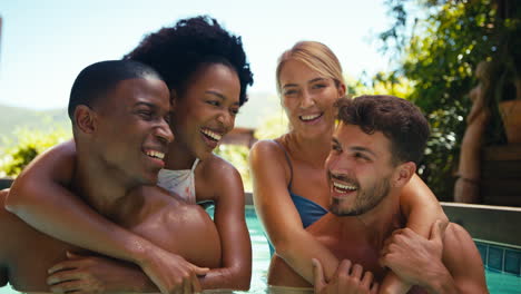 Portrait-Of-Multi-Cultural-Friends-On-Holiday-In-Swimming-Pool-With-Men-Giving-Women-Piggybacks