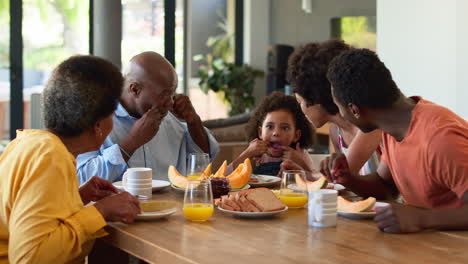 Family-Shot-With-Grandparents-Parents-And-Granddaughter-Pulling-Faces-At-Breakfast-Around-Table-At-Home