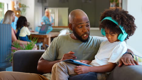 Vater-Und-Sohn-Nutzen-Zu-Hause-Ein-Digitales-Tablet,-Im-Hintergrund-Eine-Familie-Mit-Mehreren-Generationen