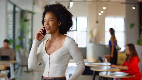 Smiling-Businesswoman-Standing-In-Busy-Open-Plan-Office-Talking-On-Mobile-Phone
