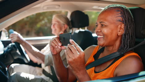 Passenger-Taking-Photo-On-Mobile-Phone-As-Two-Senior-Female-Friends-Enjoy-Day-Trip-Out-In-Car