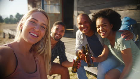 POV-Shot-Of-Friends-On-Vacation-Sitting-On-Porch-Of-Countryside-Cabin-Drinking-Beer-And-Taking-Selfie