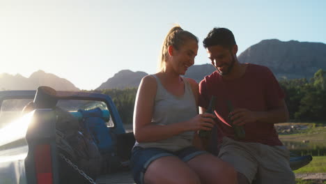 Couple-With-Backpacks-Sitting-On-Tailgate-Of-Pick-Up-Truck-On-Road-Trip-By-Lake-Drinking-Beer